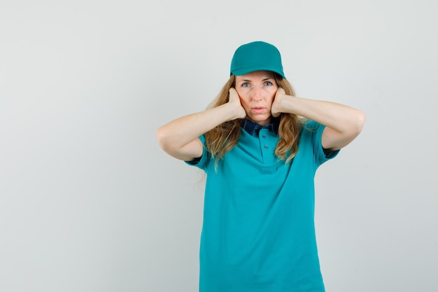 Женщина-доставщик в футболке, кепке держит уши руками и выглядит испуганной