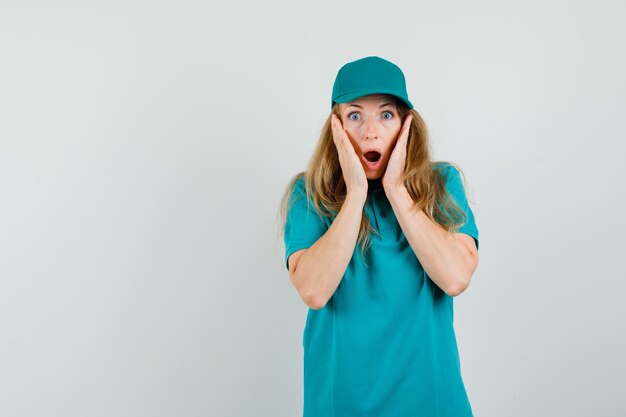 Delivery woman in t-shirt, cap holding hands near open mouth and looking shocked 