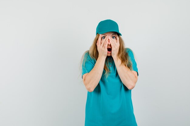 Delivery woman in t-shirt, cap looking through fingers and looking surprised 