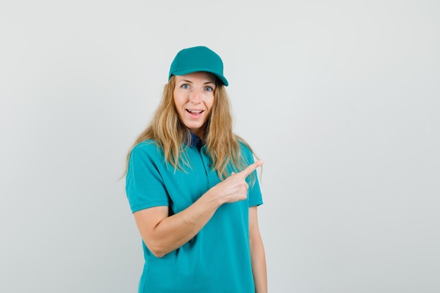 Delivery woman in t-shirt, cap pointing away and looking cheerful 