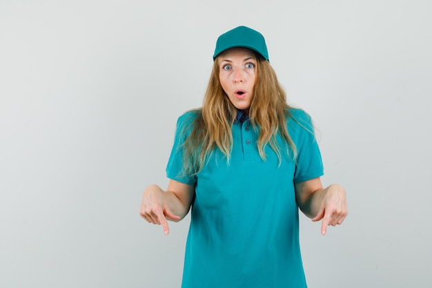 Free photo delivery woman in t-shirt, cap pointing down and looking wondered