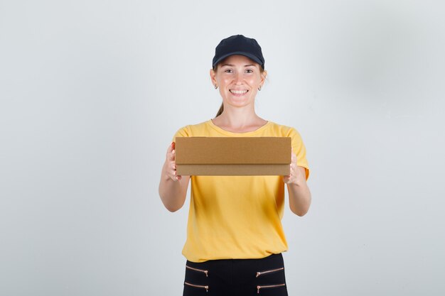 Delivery woman in t-shirt, pants, cap holding cardboard box and smiling