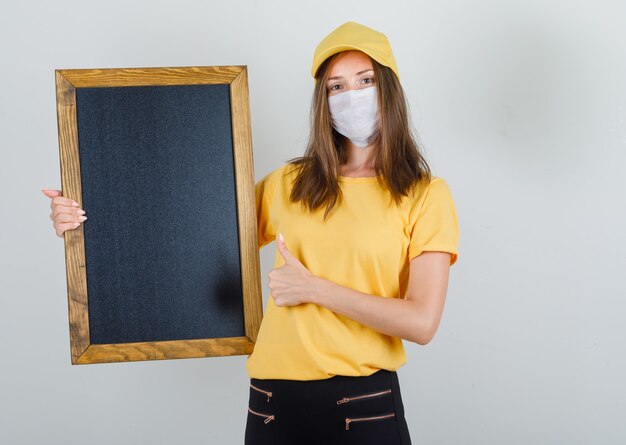 Delivery woman in t-shirt, pants, cap, mask holding blackboard with thumb up