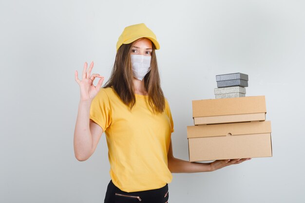 Delivery woman in t-shirt, pants, cap, mask holding boxes with ok sign and looking cheery