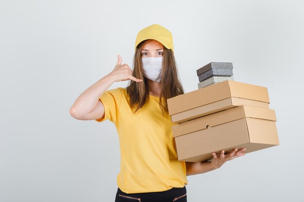 Delivery woman in t-shirt, pants, cap, mask holding boxes with phone gesture