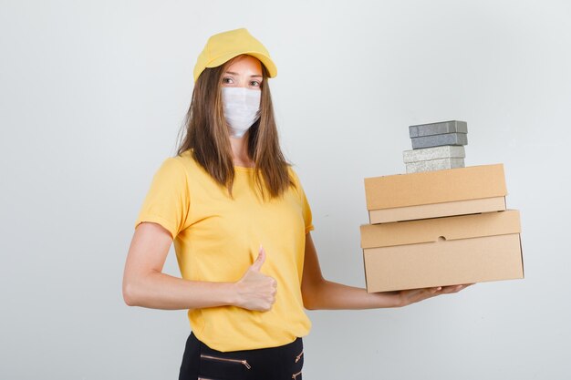 Delivery woman in t-shirt, pants, cap, mask holding boxes with thumb up and looking cheerful