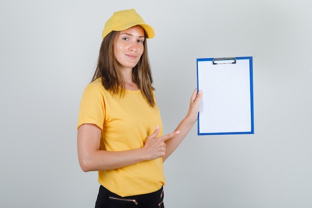Delivery woman in t-shirt, pants, cap pointing finger at clipboard and looking cheery