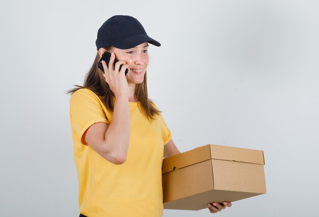 Женщина-доставщик разговаривает по смартфону с картонной коробкой в футболке, брюках и кепке