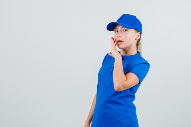 Доставщица рассказывает секрет рукой возле рта в синей футболке и кепке