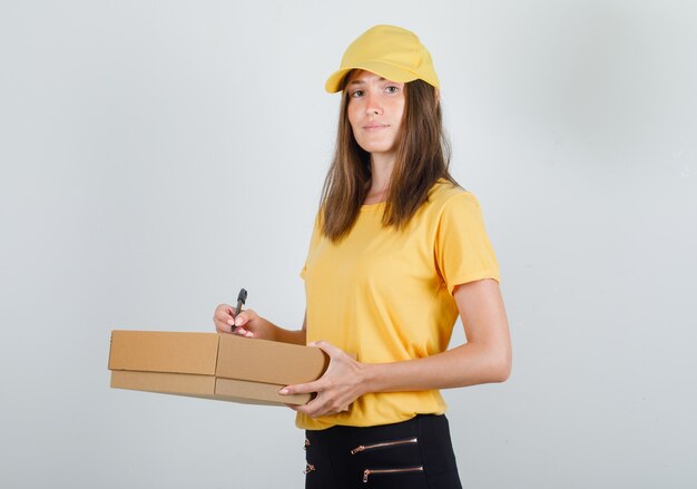 Delivery woman writing on cardboard box and smiling in t-shirt, pants and cap