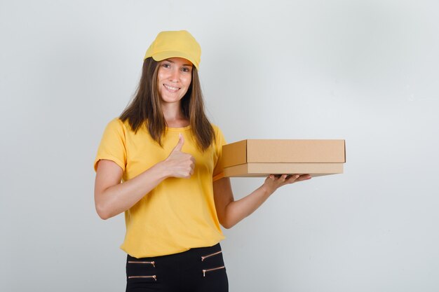 Delivery woman in yellow t-shirt, pants, cap holding cardboard box with thumb up and looking cheery