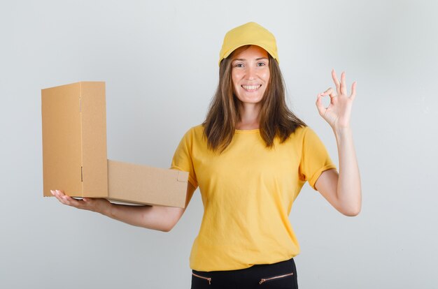 Delivery woman in yellow t-shirt, pants, cap holding opened box with ok sign and looking glad