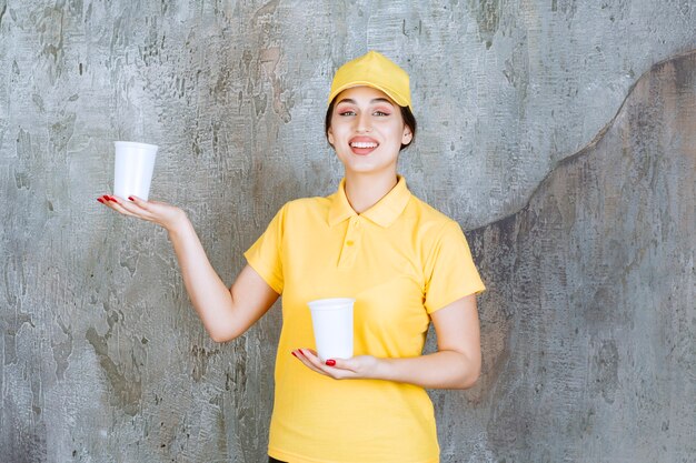 женщина-доставщик в желтой форме держит два пластиковых стаканчика с напитком и дает один другому человеку.