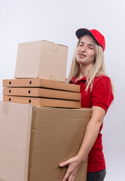 격리 된 흰 벽에 많은 상자를 들고 빨간 티셔츠와 모자를 입고 배달 젊은 여자