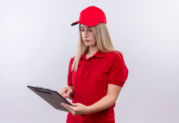 Delivery young woman wearing red t-shirt and cap is reading something on clipboard on isolated white wall