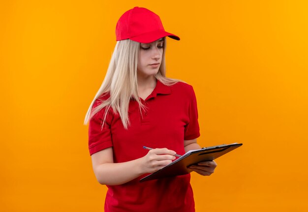 赤いTシャツとキャップを身に着けている配達若い女性は、孤立したオレンジ色の壁のクリップボードに何かを書いた