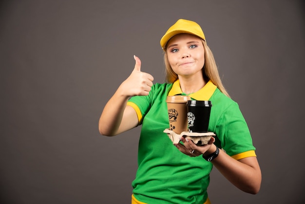 Deliverywoman holding cups of coffee and showing thumb up.