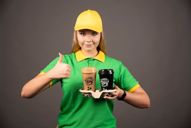Deliverywoman holding cups of coffee and showing thumb up.