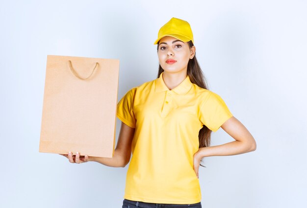 Deliverywoman in yellow cap holding brown craft paper and posing on white.