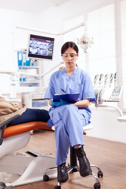 年配の女性の健康診断中に歯科医院でクリップボードにメモを取る歯科助手。歯の問題について口腔病学オフィスの医療看護師と話している年配の女性。