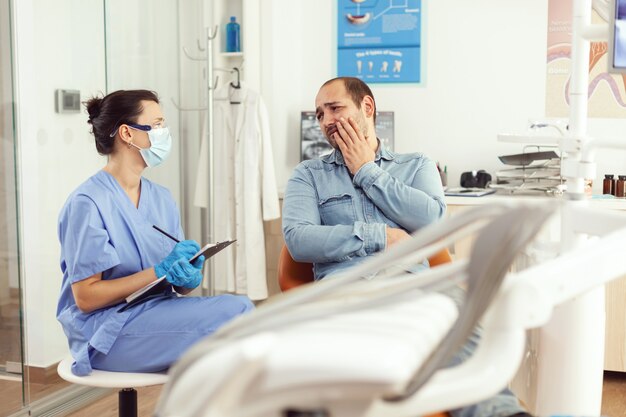 Dental assistant writing stomatology treatment on clipboard in dental clinic during sick patient check up