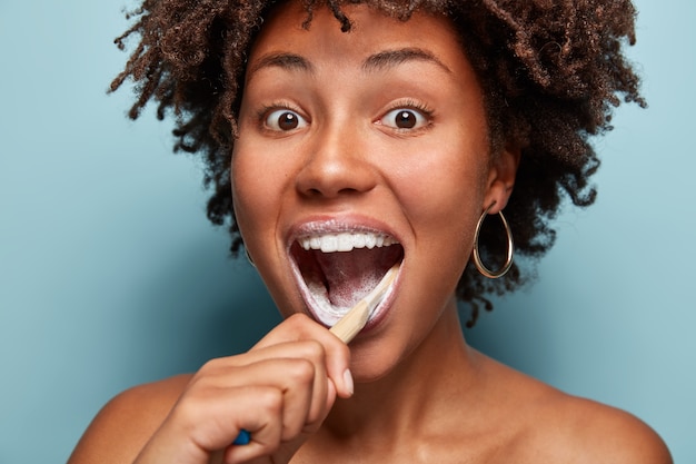 Concetto di cura, bellezza e felicità dentale. la ragazza teenager afroamericana positiva apre ampiamente la bocca, si lava i denti al mattino con spazzolino da denti e dentifricio, si sente felice, modella sul muro blu.