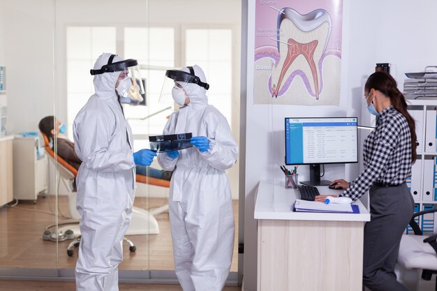 Dentist assistant discussing with doctor patient diagnosis keeping social distancing dressed in ppe suit face shiled, during global pandemic with coronavirus holding x-ray