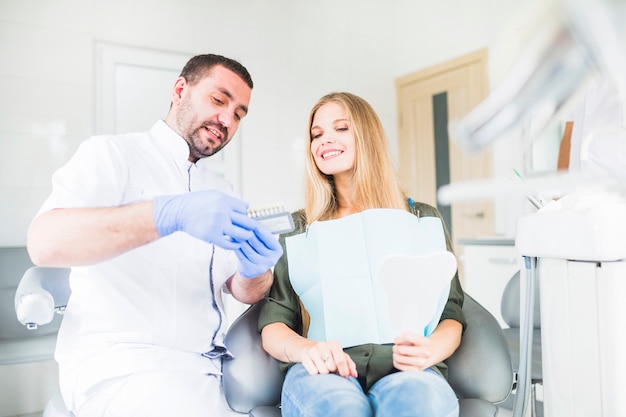 Dentist assisting happy female patient while choosing color tone of her teeth