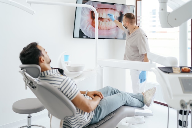 Стоматолог обсуждает с лежащим пациентом, показывая изображение его зубов на экране