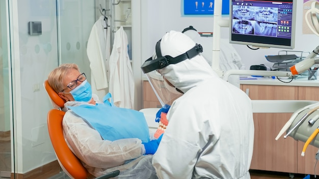 코로나바이러스 전염병 동안 치아 골격 모형을 사용하여 올바른 치과 위생을 보여주는 작업복을 입은 치과 의사. 보호복, 안면 보호 마스크, 장갑을 끼고 있는 의료 팀.