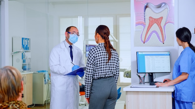 Dentist doctor interrogating woman and taking notes on clipboard standing in waiting area. Young patient explaining dental problem to stomatologist speaking in crowded reception of clinic.