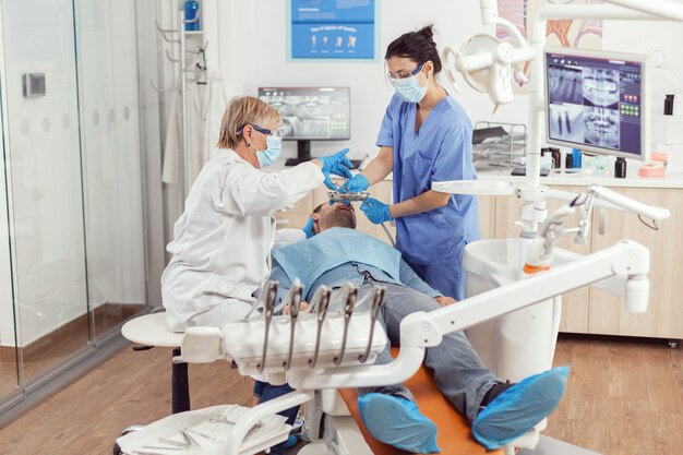 Dentist doctor and medical nurse making professional teeth cleaning to sick man patient during stomatological examination in dental office. Hospital team examining toothache preparing tooth treatment