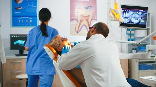 Dentist doctor working in dental unit with nurse and senior patient. Orthodontist speaking to woman sitting on stomatological chair while nurse preparing tools for examination in modern clinic