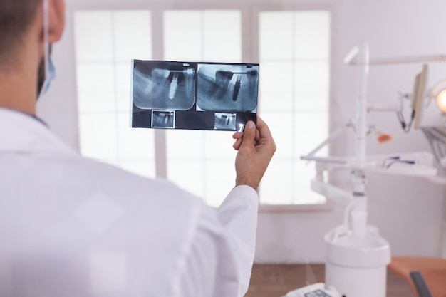 Врач стоматолог анализирует медицинскую рентгенографию ортодонтических зубов, работающих в кабинете стоматологической больницы. На заднем плане пустой кабинет ортодонта готовится к хирургической операции по уходу за зубами