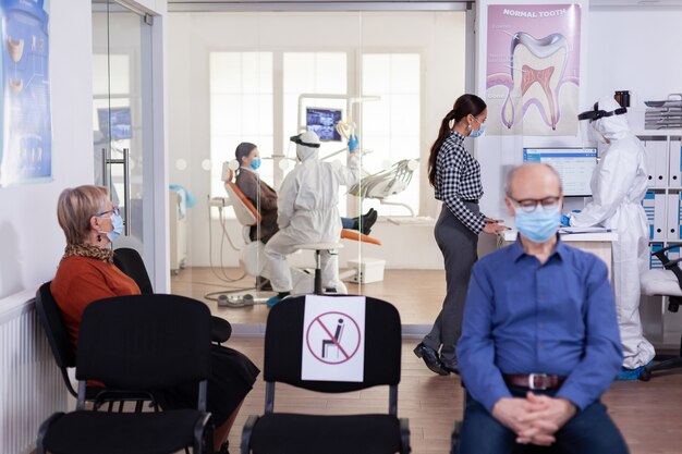 Dentist nurse dressed in ppe suit with face shiled discussing with patient in stomatology waiting room