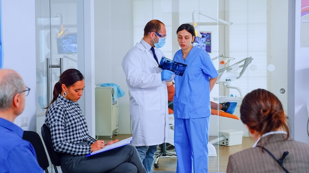 Dentist showing teeth x-ray reviewing it with nusre. Doctor and assistant working in modern crowded stomatological clinic, patients sitting on chairs in reception filling in dental forms and waiting