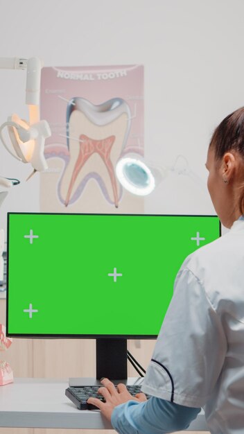 Стоматолог с помощью компьютера с горизонтальным зеленым экраном на дисплее. Специалист смотрит на монитор с цветным ключом и изолированным шаблоном для ухода за полостью рта, одновременно используя стоматологическое оборудование.