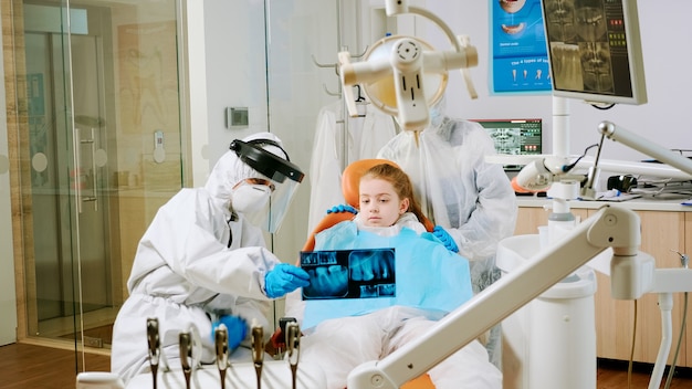 Dentist with coverall holding mouth x-ray image child patient talking with patient mother during global pandemic. Assistant and doctor talking wearing suit, coverall, protection suit, mask, gloves