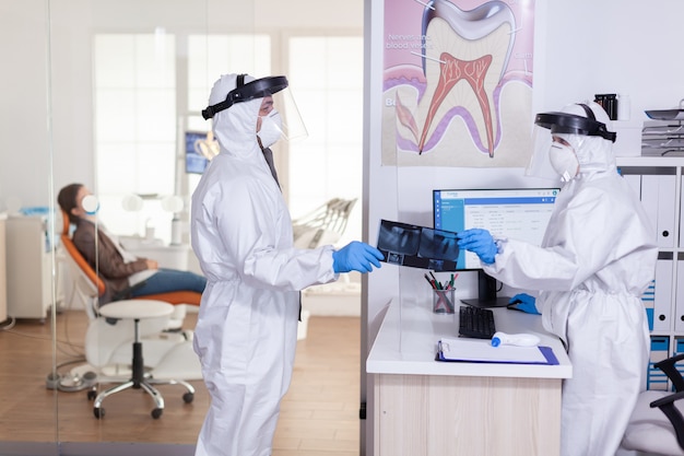 Стоматолог с защитной маской и защитным костюмом принимает рентгеновский снимок пациента от secretarty, сохраняя социальное дистанцирование во время глобальной пандемии коронавируса, область медицины стоматологии