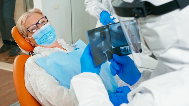 Стоматолог в маске просматривает панорамный рентгеновский снимок рта пациенту во время глобальной пандемии. Помощник и врач разговаривают с пожилой женщиной в костюме, комбинезоне, защитном костюме, маске, перчатках