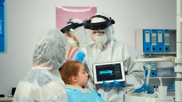Дантист в костюме ppe, указывая на цифровой экран, объясняя рентгеновский снимок матери пациентки. Медицинская бригада и пациенты в защитном комбинезоне, маске, перчатках, показывающие рентгенографию с помощью ноутбука