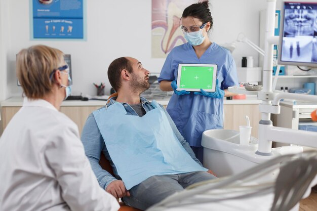 Dentist woman holding mock up green screen chroma key tablet with isolated display discussing oral hygiene with man patient during stomatological consultation in dental office. stomatology concept