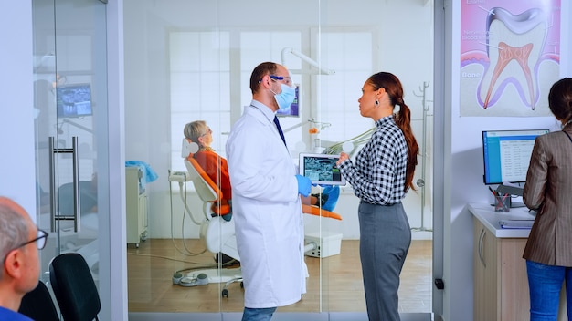 歯科医院の待機エリアに立っているタブレットを使用して患者に歯のX線写真を示す歯科医。混雑したオフィスでの治療を説明する女性と一緒に歯科X線撮影をレビューする口腔病学者