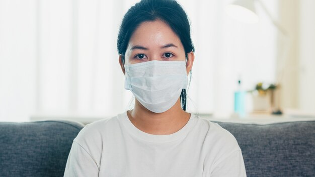 落ち込んでいるアジアビジネスの女性の社会的距離が家にいるとき、家のリビングルームのソファーに座っている防護マスクと自己検疫時間、中国でのパンデミック、コロナウイルスの概念。