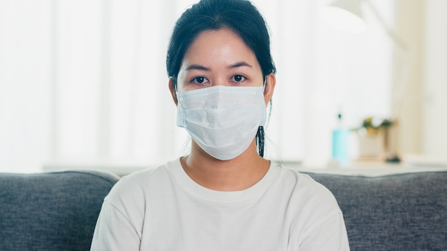 Подавленная азиатская бизнес-леди нося защитную маску сидя на софе в живущей комнате на доме когда социальное дистанцирующ пребывание дома и время карантина собственной личности, пандемия в фарфоре, концепции коронавируса.