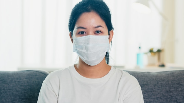 Depressed Asian business woman wearing protective mask sitting on sofa in living room at house when social distancing stay at home and self quarantine time, pandemic in china, coronavirus concept.