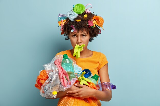 Depressed black woman with crisp hair, picks up garbage, has annoyed negative facial expression, cleans environment isolated over blue wall, sorts litter. People, recycling, volunteering concept