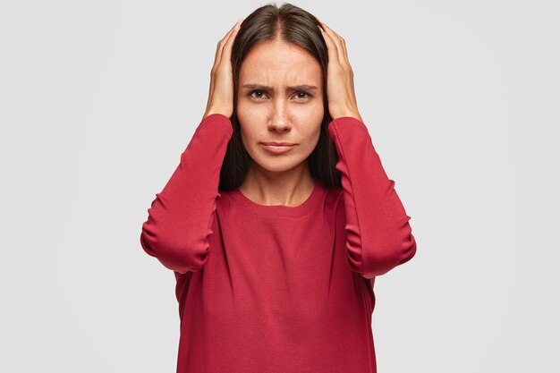 Depressed brunette young European woman has stressful facial expression, keeps hands on head