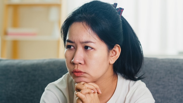 Depressed crying Asian woman stressed with headache sitting on sofa in living room at house