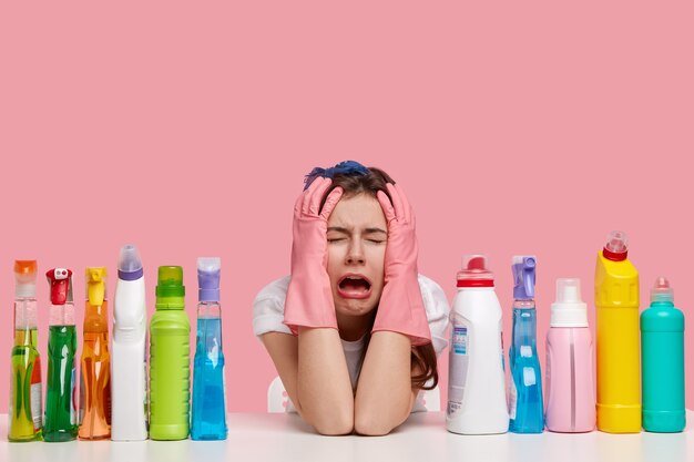 Depressed crying woman looks stressfully, keeps both hands on head, wears protective gloves